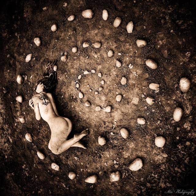 Photo art by Niiv Photography: a woman lying curled on the bare earth within a spiral of stones, seen from above in warm sepia tones