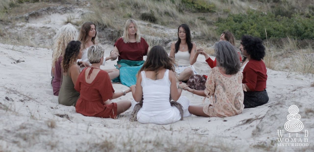 Women holding hands in a circle on a sandy dune, eyes closed, dressed in white and rust red.