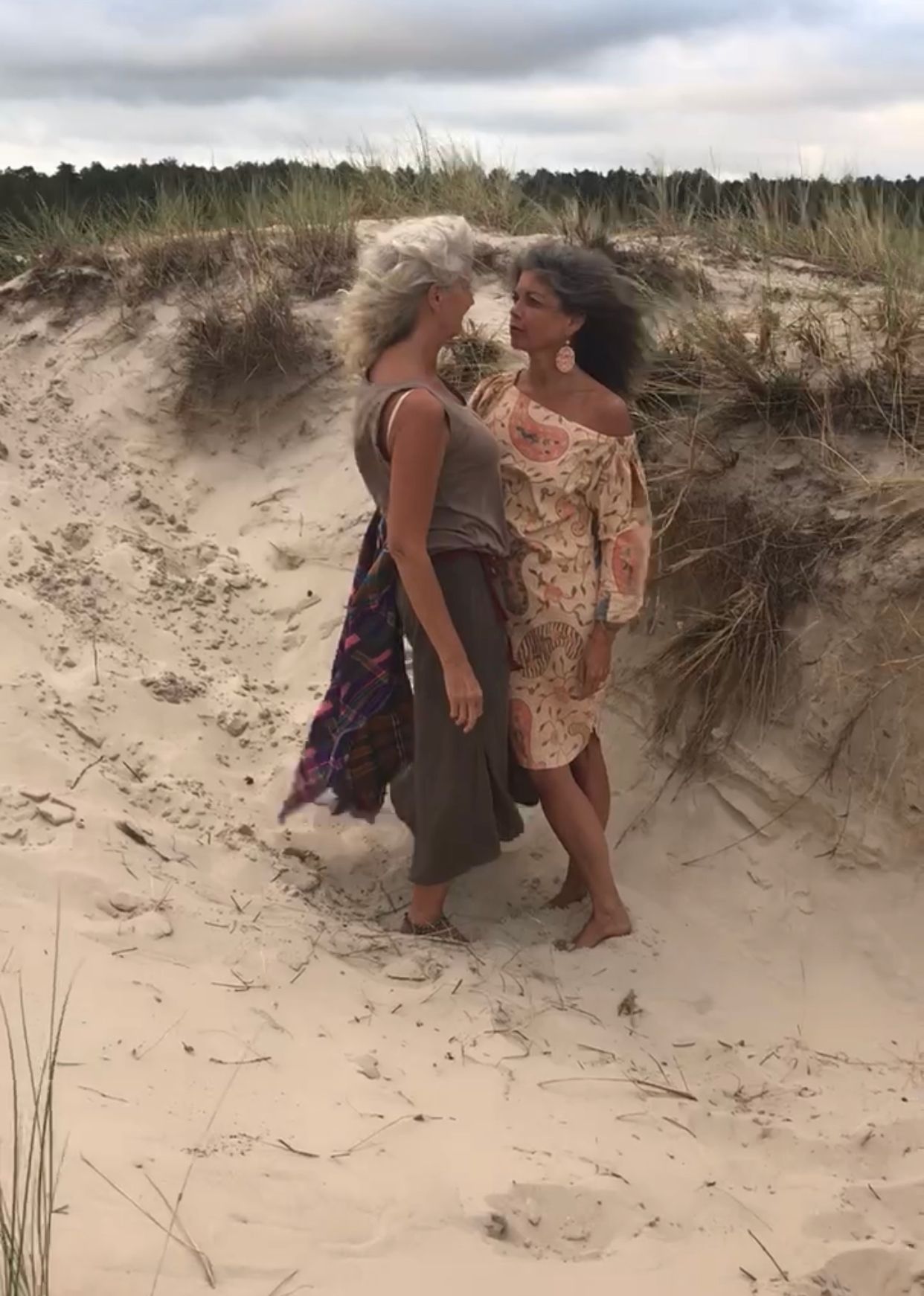 Two women with windswept grey hair stand face to face between grassy dunes.