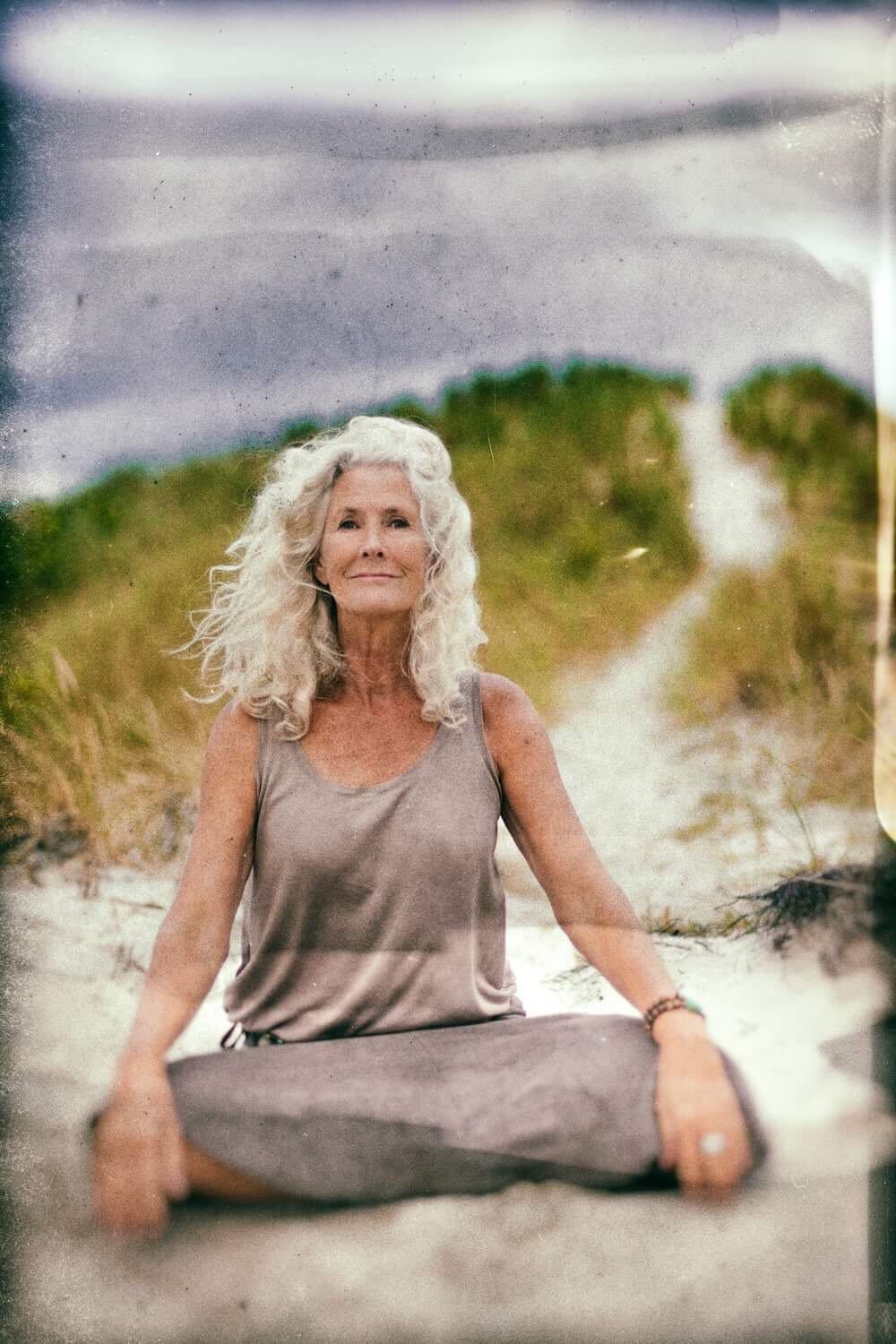A woman with long white curly hair sitting cross-legged on a dune path