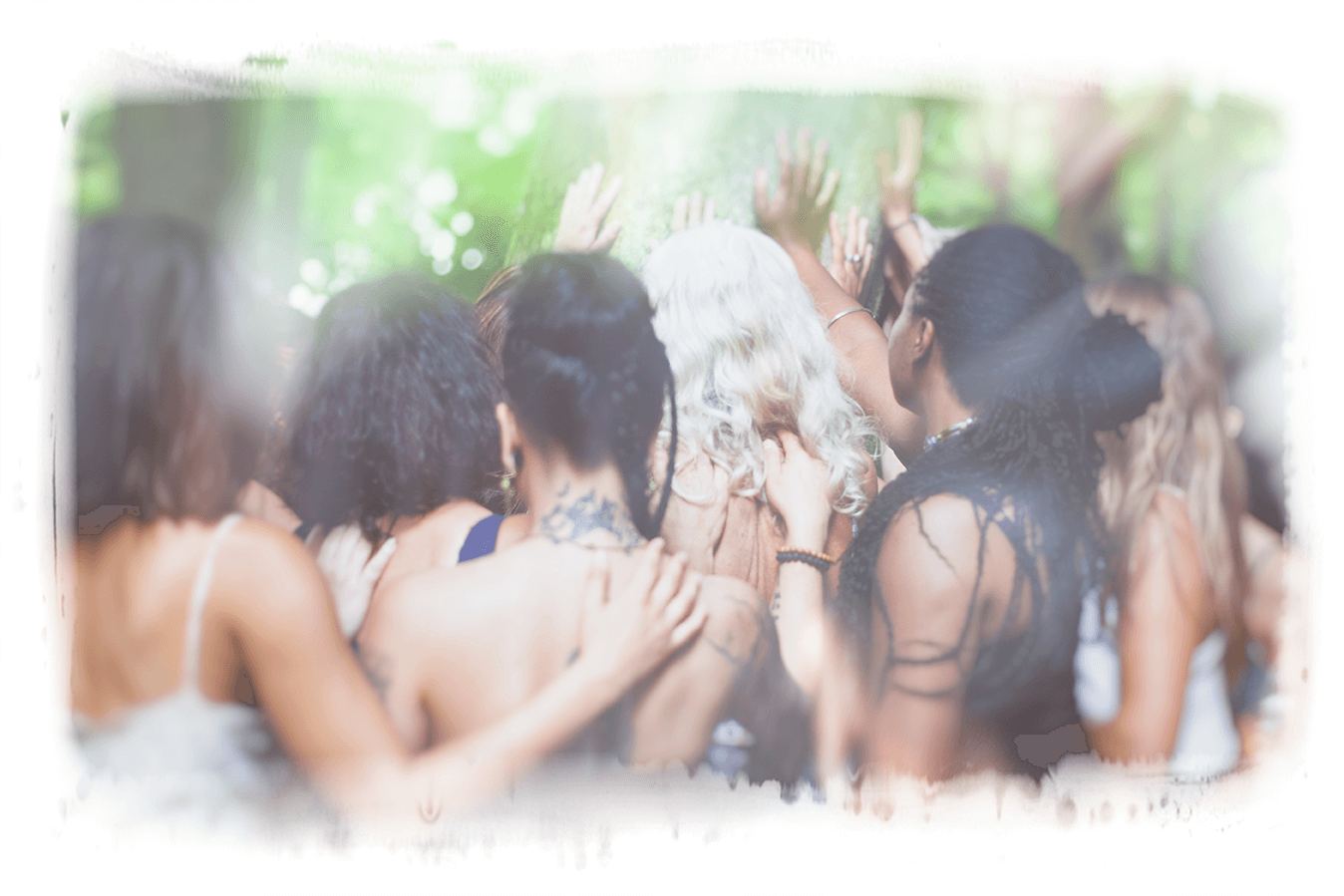 A group of women standing close together with their arms around one another, hands raised toward the sunlit canopy of a green forest.