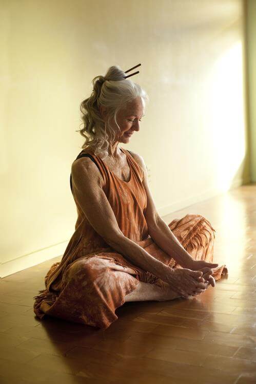 A silver-haired woman in a terracotta dress sitting in meditation on a wooden floor