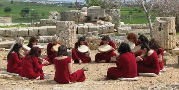 Women in red dresses sitting in a circle with frame drums among ancient stone ruins.