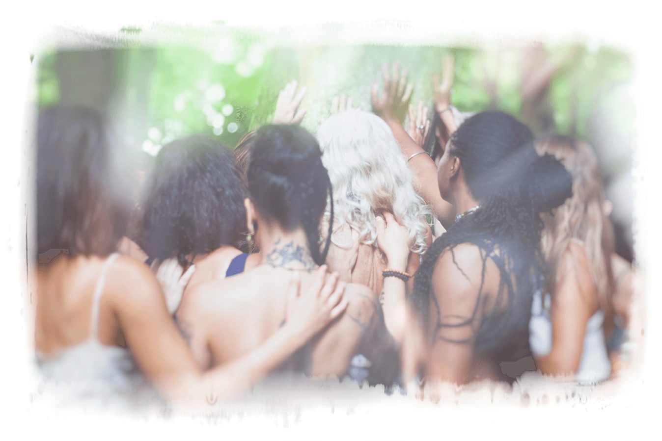 Women with their arms around each other, raising their hands toward a sunlit green forest.