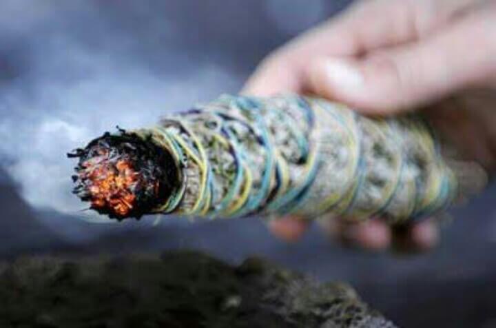 A hand holding a smouldering bundle of dried white sage, bound with coloured thread