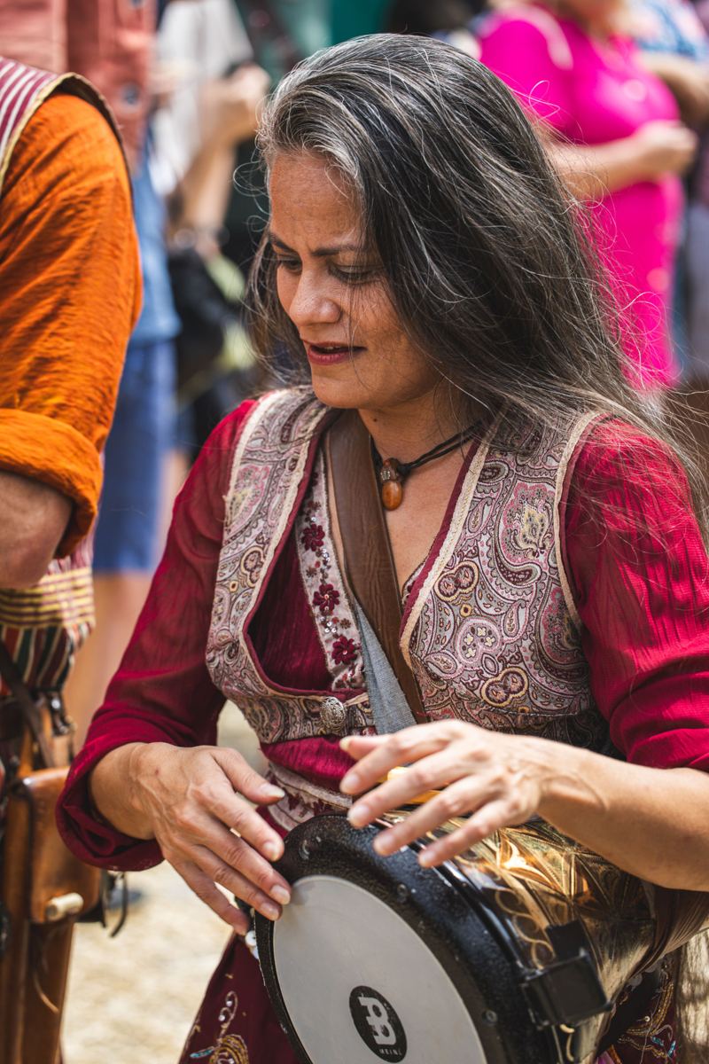 A grey-haired woman in a red embroidered dress plays a hand drum at a crowded gathering.