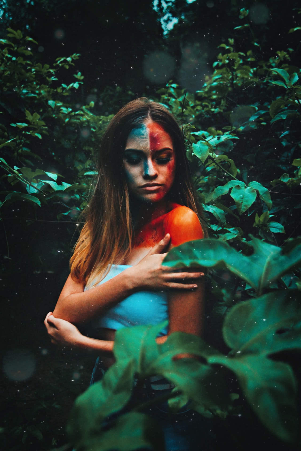 A woman with half her face painted red standing among dark green leaves, one hand on her shoulder.
