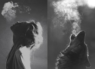 A woman and a howling wolf side by side in black and white, their breath rising as mist into the cold air.