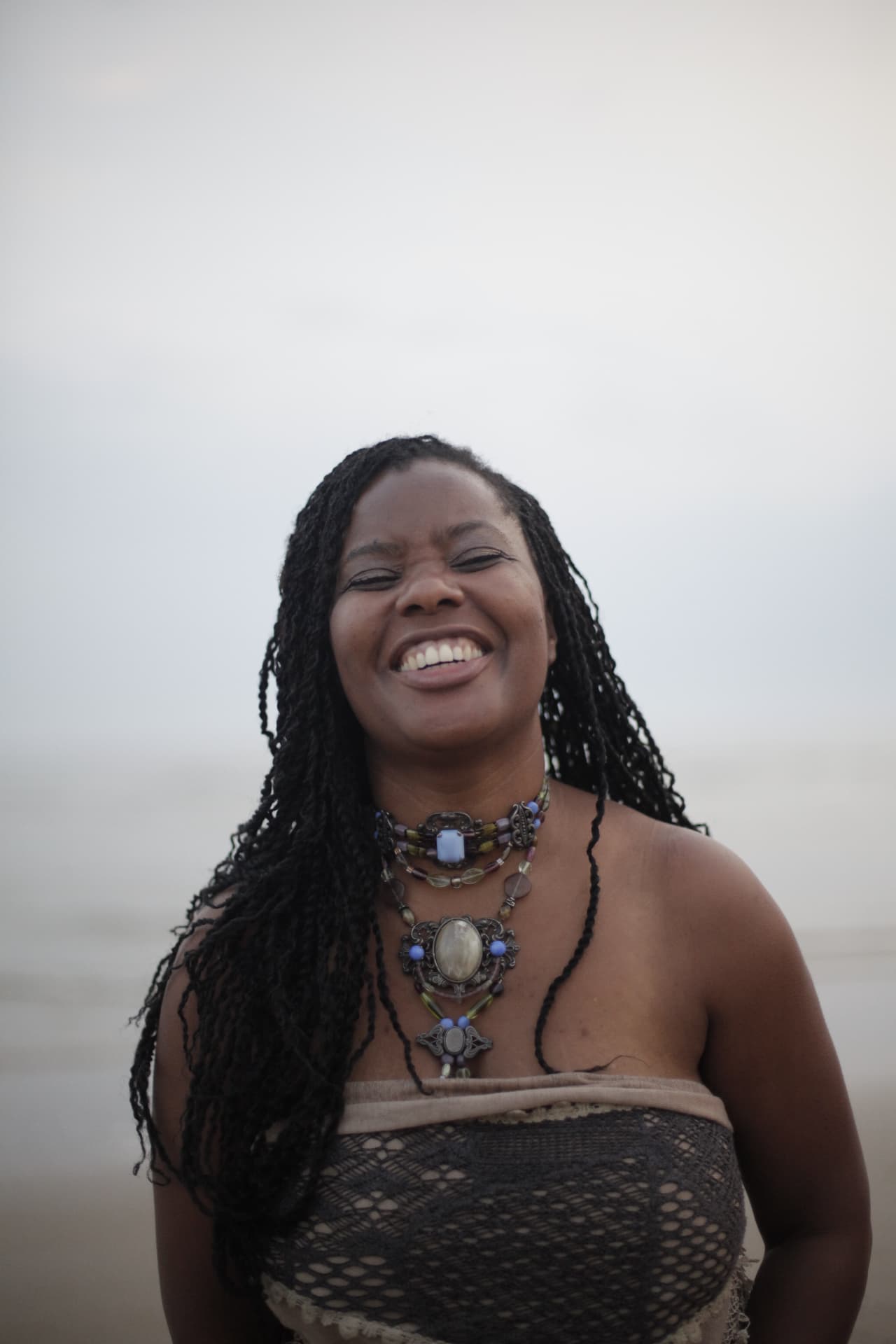 A smiling woman with long braids and layered necklaces outdoors
